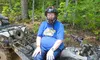 A person wearing a helmet and blue shirt is sitting on a muddy ATV in a wooded area.
