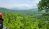 A person wearing a red helmet sits in an off-road vehicle, overlooking a lush, green forest under a clear blue sky with clouds.