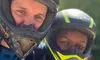 Two people wearing helmets are smiling and embracing while outdoors in a sunny setting.