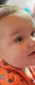 A baby wearing an orange, patterned shirt is looking to the side with a curious expression.