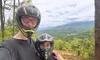 Two people in helmets are posing for a selfie with a scenic, mountainous landscape in the background.