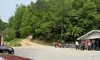 The image shows a group of people wearing helmets near a building with ATVs parked beside them, surrounded by a forested area and gravel path, with a red car in the foreground.