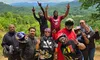 A group of people in outdoor gear pose enthusiastically with peace signs and gestures in a scenic mountainous area.