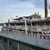 A decorated riverboat with patriotic bunting is docked alongside a river under a partly cloudy sky.