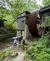 Three people are standing near a large wooden waterwheel and rustic building in a forested area by a stream.