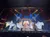 Dancers in flowing white costumes perform energetically on a festive, decorated stage.