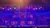The image shows an indoor stage set with a vibrant, colorful backdrop resembling a palace, lit by hanging lanterns, with an audience gathered below.