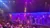 The image shows a theater with an exotic, Middle Eastern-style stage set under vibrant purple lighting, filled with an audience seated and standing.