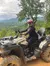 A person wearing a helmet and protective gear is sitting on an ATV in a mountainous, forested area.