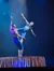 Two acrobats perform a balancing act on a platform, with one person supporting the other by the waist, under dramatic blue lighting.