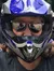 A person wearing a helmet and sunglasses is sitting on a muddy Kawasaki ATV near a building and a fence.