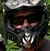 A couple wearing helmets stands together in a wooded area with ATVs nearby.