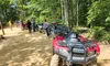 A group of people are gathered near a line of parked ATVs on a dirt path in a forested area.