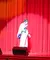 A performer in a glittery costume sings into a microphone on stage in front of red curtains.