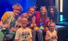 A group of performers poses cheerfully with two young children in front of a stage with colorful lights.