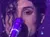 A person in a shiny silver jacket is singing into a microphone on a dimly lit stage.