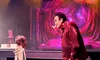 A man in a red shirt is engaging with a young boy in the audience on a stage with dramatic lighting and red curtains in the background.