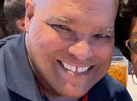 A smiling couple is seated at a dining table, with dessert plates in front of them, including a slice of cake in a takeaway container.