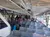 A group of people is seated on a boat with rows of metal chairs, some facing forward and others interacting or looking around.