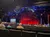 The image depicts an audience seated in a theater with a stage illuminated in red, displaying