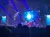 The image captures a lively stage performance with vibrant lighting, dancers, and a large, illuminated backdrop, viewed by an audience.