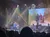 A band performs energetically on stage with a large video screen behind them showing two people riding on an off-road vehicle.