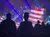 The image shows a vibrant stage with a large American flag backdrop and silhouetted people in the foreground, under colorful spotlights.