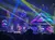 A vibrant stage performance is taking place with colorful lights and performers in dynamic outfits, set against a backdrop of neon triangular designs.