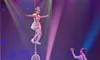A performer balances on a unicycle perched on a large ball, while another holds up a ring, with colorful stage lights illuminating the scene.