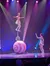A performer balances on a unicycle perched on a large ball, while another holds up a ring, with colorful stage lights illuminating the scene.