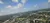 The image shows an aerial view of a cityscape with a highway, green areas, and scattered clouds in a bright blue sky.