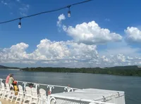 A sunny day on a riverboat with people sitting on deck chairs, overlooking a scenic landscape and string lights above.