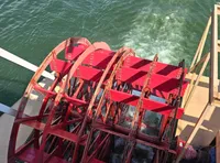 The image shows a large red paddlewheel on a boat splashing through greenish water.