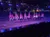 A group of people on horseback are performing in an arena, carrying American flags and wearing illuminated costumes, under vibrant stage lighting with an audience watching.