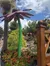 The image features a large, colorful flower sculpture in a garden setting, with blooming plants, a small stream, and people walking in the background under a blue sky with fluffy clouds.