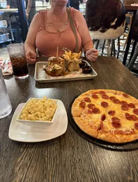 A person is seated at a table with a tray of food including a burger and fries, while a pepperoni pizza and a dish of macaroni and cheese are placed on the table.