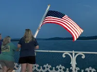 Three people are standing on a boat looking at the water, with an American flag waving prominently in the foreground.