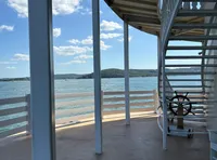 The image shows an open deck of a boat with a spiral staircase and a ship's wheel, overlooking a body of water and distant hills under a blue sky with clouds.