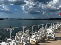 White Adirondack chairs are lined up on a sunny deck overlooking a body of water with a backdrop of dramatic clouds in the sky.