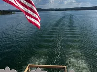 A view from a boat with an American flag fluttering in the breeze, overlooking a calm body of water under a partly cloudy sky.