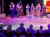 A group of performers is entertaining an audience on stage, with some musicians playing instruments and others standing in front of a seated crowd.