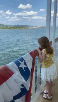 A young girl in a yellow floral dress is gazing at the water from a boat deck, holding onto a railing draped with a decorative banner featuring stars.