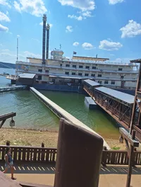A large, vintage-style riverboat docked at a pier on a sunny day, with a person walking along a boardwalk in the foreground.