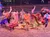 A group of performers in colorful costumes dance energetically around a small fire in an arena, with an audience watching from tiered seating in the background.