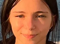A person is standing on a balcony overlooking a body of water with a tree-lined shore in the background.