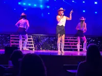 Three performers in cowboy hats and boots are on stage, with one singing into a microphone under vibrant blue lights.