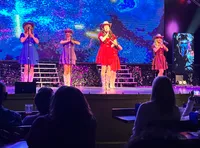 Four performers in colorful dresses and cowboy hats sing on a stage with a vibrant, abstract background.