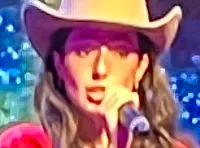 Four performers in cowboy hats and dresses are singing on a brightly lit stage, with a large digital crown displayed behind them.