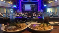 The image shows a theater setting with a stage lit in purple, plates of steak and potatoes on a table in the foreground, and an audience seated in the background.