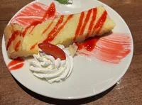 A slice of cheesecake drizzled with red sauce is served on a white plate with whipped cream, a strawberry slice, and a mint leaf garnish.